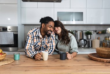 Multi-ethnic couple standing with tea cups in the kitchen and looks at each other with a love. African black man and a caucasian woman enjoy morning coffee together at home talking about life