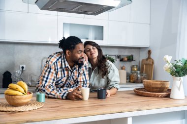 Multi-ethnic couple standing with tea cups in the kitchen and looks at each other with a love. African black man and a caucasian woman enjoy morning coffee together at home talking about life