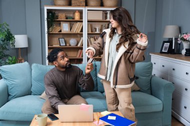 Young woman asking for credit card from her husband to go to shopping while he working from home on laptop. Freelance male unwillingly giving his bank card to his wife to spend their money on buying