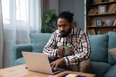 Young freelance business man or exchange student working remotely from his home office on laptop computer, having issue with internet connection or web page. Nervous African American work problems