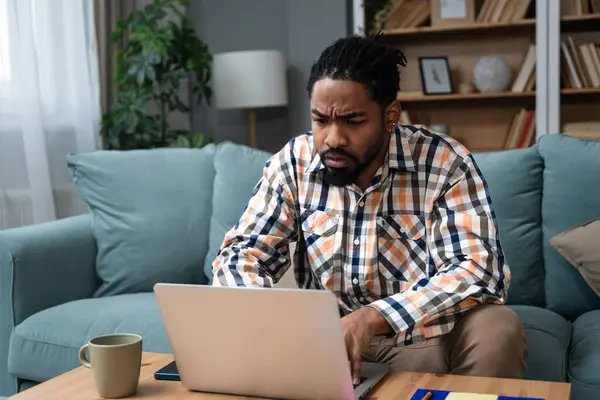 Young freelance business man or exchange student working remotely from his home office on laptop computer, having issue with internet connection or web page. Nervous African American work problems