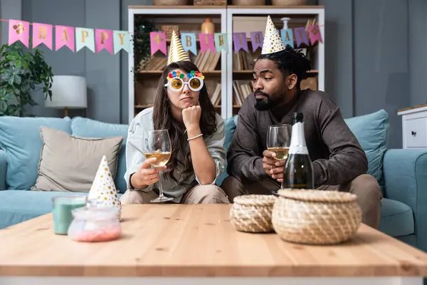 Two young people sitting on sofa with birthday caps, sad because no one came to celebrate birthday and party in their apartment. Disappointed man and woman boyfriend and girlfriend without friends