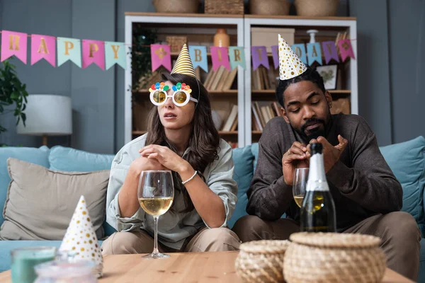Two young people sitting on sofa with birthday caps, sad because no one came to celebrate birthday and party in their apartment. Disappointed man and woman boyfriend and girlfriend without friends