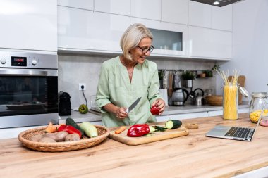 Üst düzey bir dul ev mutfağındaki evinde sebzelerden vejetaryen sağlıklı yiyecekler pişiriyor, bilgisayarını online tarifler için kullanıyor, uzun zaman sonra randevusu için yemek hazırlıyor.