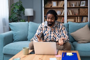 Kulaklıklı genç Afro-Amerikalı bir adam dizüstü bilgisayar ve akıllı telefon kullanarak internet üzerinden arıyor. Kanepenin üzerinde oturmuş, web kamerasındaki ana konuşuyor. İnternetteki iş ürünleri stratejisi toplantısı.