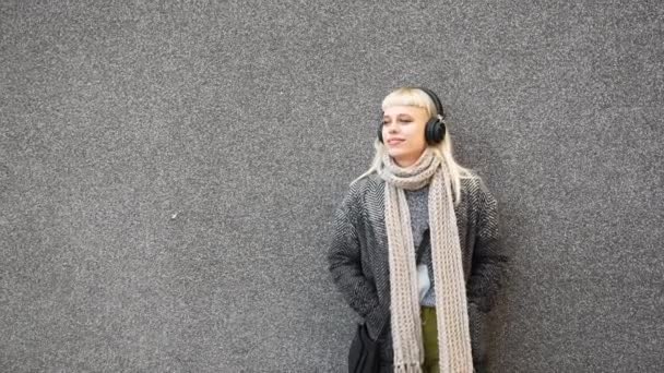 年轻可爱的嬉皮士嬉皮士女人穿着现代服装 头戴无线耳机 靠着墙壁听音乐 金发女子 头戴耳机在街上等着 — 图库视频影像