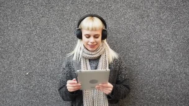 ハッピーヒッピーの女の子はタブレットをブラウズし 通りの壁に傾いているヘッドフォンで音楽を聞いています 同僚がワイヤレスヘッドセットで音楽を聴くのを待っている間 オンラインで働くヒップスター女性 — ストック動画