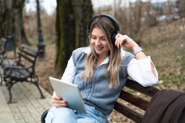 Genç iş kadını parkta oturuyor dijital tablet üzerinde çalışıyor kablosuz kulaklıkla müzik dinliyor. İşadamı serbest çalışıyor. Öğle arasında ofis işine ara veriyor.