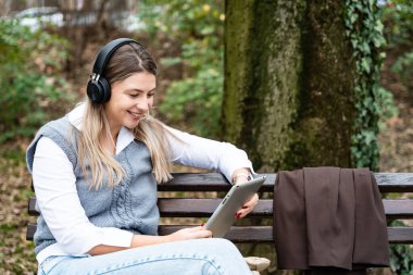 Genç iş kadını parkta oturuyor dijital tablet üzerinde çalışıyor kablosuz kulaklıkla müzik dinliyor. İşadamı serbest çalışıyor. Öğle arasında ofis işine ara veriyor.