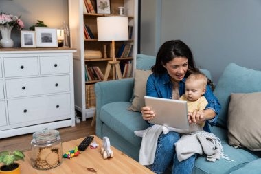Genç anne, evdeki kanepede otururken dijital tablette çizgi film ve komik videolar oynayarak yeni doğmuş bebeğini eğlendirmeye çalışıyor. İnterneti küçük çocuğuyla eğlenmek için kullanan bir kadın..