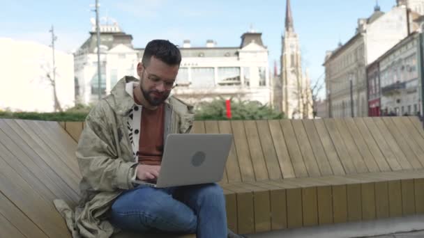 年轻的商人坐在写字楼外的长椅上 带着笔记本电脑上网工作 独立的国外自由职业嬉皮士计算机程序员专家 旅行时编程 — 图库视频影像