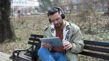 Genç bağımsız stil sahibi hippi işadamı parkta oturmuş dijital tablet üzerinde çalışıyor. Bir iş adamı iş için kablosuz teknolojiyi kullanıyor, ofis ve gürültüden dinleniyor.