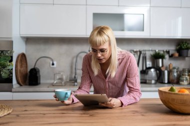 Genç iş kadını, küçük bir şirket sahibi, girişimci iş adamı pazarlama ve ürün stratejisi geliştiricisi, mutfakta ilk kahveyi istatistik ve e-postaları görmek için dijital tablet kullanarak içiyor.