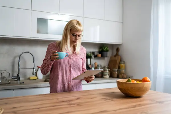 Young business woman, small company owner, startup businessperson expert marketing and product strategy developer, drinking first coffee in the kitchen using digital tablet to see statistics and mails