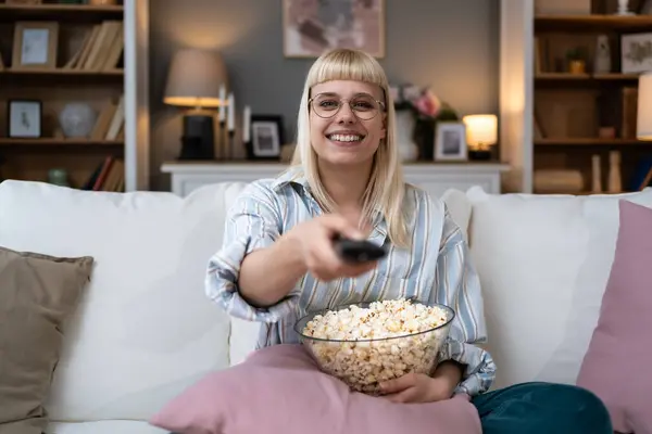 Young happy woman changing channels with remote control while watching TV and eating popcorn in the evening at home. Relaxed hipster female enjoying at weekend lazy day