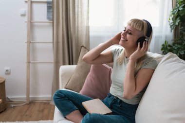 Evdeki kanepede dinlenen genç bir kadın. Kablosuz kulaklıklardan müzik dinlemek ve kitap okumak. Kitap seven kadın, rahat dairesinde dinlenmek için boş zaman ve hafta sonu aktivitelerinin tadını çıkarıyor.