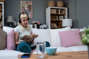 Evdeki kanepede dinlenen genç bir kadın. Kablosuz kulaklıklardan müzik dinlemek ve kitap okumak. Kitap seven kadın, rahat dairesinde dinlenmek için boş zaman ve hafta sonu aktivitelerinin tadını çıkarıyor.