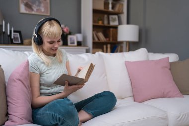 Evdeki kanepede dinlenen genç bir kadın. Kablosuz kulaklıklardan müzik dinlemek ve kitap okumak. Kitap seven kadın, rahat dairesinde dinlenmek için boş zaman ve hafta sonu aktivitelerinin tadını çıkarıyor.