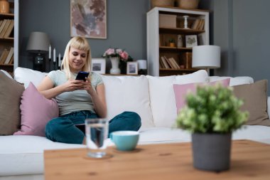 Mutlu bin yıllık sarışın kız sosyal medyayı kontrol ediyor evde akıllı telefon kullanıyor gülümseyen genç kadın cep telefonu kullanıyor oyun alışverişi yapıyor internetten teslimat siparişi veriyor kanepede buluşuyor.