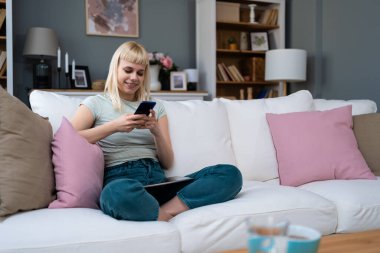 Mutlu bin yıllık sarışın kız sosyal medyayı kontrol ediyor evde akıllı telefon kullanıyor gülümseyen genç kadın cep telefonu kullanıyor oyun alışverişi yapıyor internetten teslimat siparişi veriyor kanepede buluşuyor.