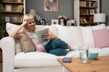 Mutlu bin yıllık sarışın kız elinde dijital tabletle sosyal medyayı kontrol ediyor gülümseyen genç kadın internet üzerinden oyun alışverişi yapıyor kanepede rahatla sipariş veriyor. Online randevu