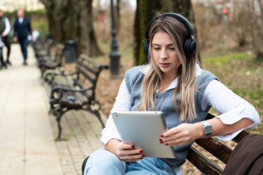 Genç başarılı iş kadını bankta oturmuş dijital tablet üzerinde çalışıyor ve kablosuz kulaklıkla podcast dinliyor. Ofis binası dışında profesyonel kadın ofis çalışanı araştırması.