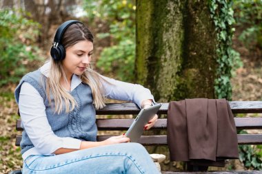 Genç başarılı iş kadını bankta oturmuş dijital tablet üzerinde çalışıyor ve kablosuz kulaklıkla podcast dinliyor. Ofis binası dışında profesyonel kadın ofis çalışanı araştırması.