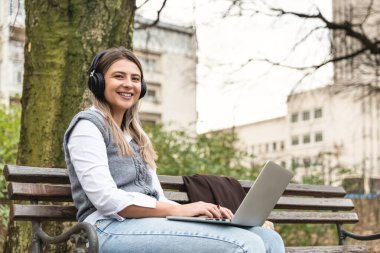 Genç başarılı iş kadını bankta oturuyor dizüstü bilgisayarda çalışıyor ve kablosuz kulaklıkla podcast dinliyor. Ofis binası dışında profesyonel kadın ofis çalışanı araştırması.