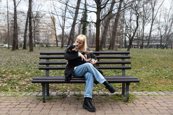 Young generation Z hipster girl sitting in park on a bench with her adopted dog, whom she rescued from the street from starvation, and gives him love and attention. Animal lover enjoying with her pet.