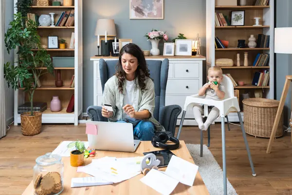 Online ödemeler. Oğlu ya da kızı olan mutlu genç bir anne dizüstü bilgisayar ve kredi kartı kullanıyor, sevimli bir çocukla internet alışverişi yapıyor. Bekar anne internette bebek alışverişi yapıyor.