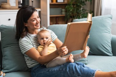 Young happy mother reading a fairytale story book to her baby. Mommy and kid sitting on sofa at home enjoying in imagination. Parent and child lifestyle. clipart