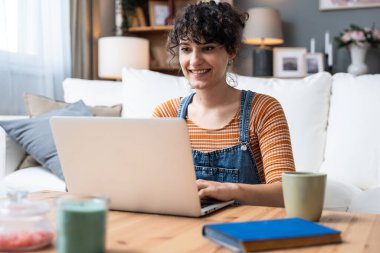 Evde bilgisayarla çalışan mutlu ve serbest bir genç kadın. Çevrimiçi öğrenim gören çekici iş kadını, dizüstü bilgisayar, web sörfü bilgileri veya internet mağazasından alışveriş yapıyor..