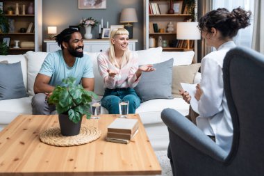 İlişkilerindeki sorunları çözmeye çalışan iki kişi, genç ve çeşitli bir çift. Ofiste terapistin önünde konuşurken gülümseyen çok ırklı eşler. Evlilik terapisi. Aile uzlaşması.