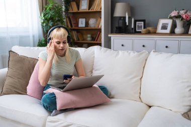 Çekici genç bir kadın evinde görüntülü konuşma yapmak için dizüstü bilgisayarını ve kulaklıklarını kullanarak kanepede bacak bacak üstüne atmış oturuyor. Üniversiteli kız online randevu, müzik dinlemek, evde çalışmak.