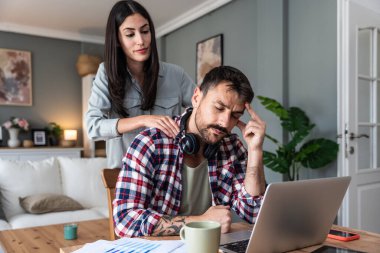 Genç ve stresli bir adam dizüstü bilgisayarda çalışıyor ve kadın stresi azaltmak için ona masaj yapıyor. Erkek işadamı omuz ağrısı çekiyor, kadın ona masaj yapıyor.