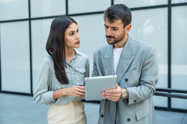 Dijital tablet kullanan ve dışarıda dikilen iki iş arkadaşı. Modern iş arkadaşları işe giderken ofis binasının önünde sohbet ediyorlar. İş adamları işbirliği.