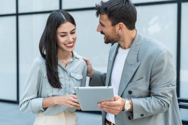 Dijital tablet kullanan ve dışarıda dikilen iki iş arkadaşı. Modern iş arkadaşları işe giderken ofis binasının önünde sohbet ediyorlar. İş adamları işbirliği.