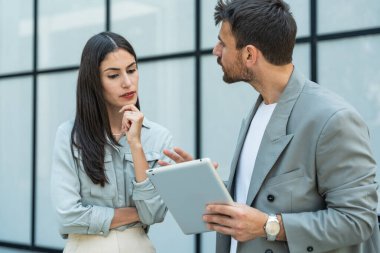 Dijital tablet kullanan ve dışarıda dikilen iki iş arkadaşı. Modern iş arkadaşları işe giderken ofis binasının önünde sohbet ediyorlar. İş adamları işbirliği.