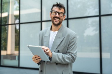 Dijital tabletle ofis binasının önünde duran genç bir iş adamı takım lideri. Finansal bölgedeki iş yerinin dışında başarılı bir tasarımcının portresi. İş insanları