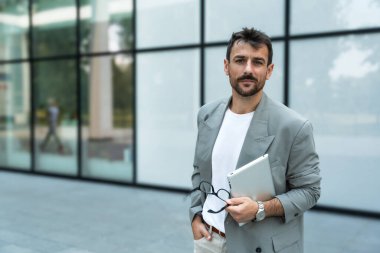Dijital tabletle ofis binasının önünde duran genç bir iş adamı takım lideri. Finansal bölgedeki iş yerinin dışında başarılı bir tasarımcının portresi. İş insanları