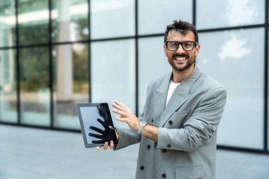 Dijital tabletle ofis binasının önünde duran genç bir iş adamı takım lideri. Finansal bölgedeki iş yerinin dışında başarılı bir tasarımcının portresi. İş insanları