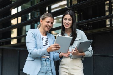 İki iş kadını bilgisayar ve dijital tabletle ofis binası dışında. Modern iş hayatında kullanılan teknoloji. Sürdürülebilir enerji uzmanları, geliştiriciler ve tasarım uzmanları birlikte çalışıyorlar.