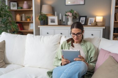 Rahat genç beyaz kadın modern cihaz okuyucu üzerine bir kitap okudu. Evdeki koltukta rahatça oturuyordu. Gözlük takan ve dijital tablet bağlantısı kullanan güzel kadın portresi.