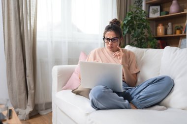 Sunny Cozy apartmanında dizüstü bilgisayarda çalışan ciddi yaratıcı bir kadın kanepenin üzerinde dinleniyor. Başarılı yaratıcı kadın girişimci uzaktan kumandalı ticari işler. Profesyonel tasarım.