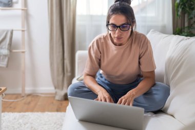 Sunny Cozy apartmanında dizüstü bilgisayarda çalışan ciddi yaratıcı bir kadın kanepenin üzerinde dinleniyor. Başarılı yaratıcı kadın girişimci uzaktan kumandalı ticari işler. Profesyonel tasarım.