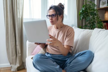 Sunny Cozy apartmanında dizüstü bilgisayarda çalışan ciddi yaratıcı bir kadın kanepenin üzerinde dinleniyor. Başarılı yaratıcı kadın girişimci uzaktan kumandalı ticari işler. Profesyonel tasarım.