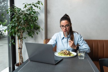 Genç iş kadını öğle arasında kafeteryada uzaktan çalışıyor. Kadın ofis çalışanı restoranda yemek yerken dizüstü bilgisayar kullanıyor ya da internetten komik içerikler izliyor..