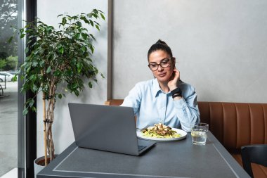 Genç iş kadını öğle arasında kafeteryada uzaktan çalışıyor. Kadın ofis çalışanı restoranda yemek yerken dizüstü bilgisayar kullanıyor ya da internetten komik içerikler izliyor..