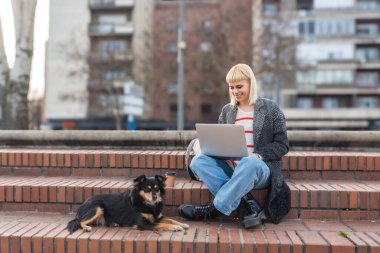 Genç yetişkin bir kadın ya da kız, üniversite öğrencisi, değişim öğrencisi, serbest çalışan ya da evcil ya da kurtarılmış köpeğiyle dışarıda laptopta çalışan bir göçmen. İş dünyası dışarıda, özgürlük kavramı