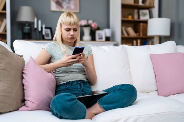 Mutlu, rahat, genç bir kadın koltukta oturmuş telefonla gülüyor, elinde akıllı telefonuyla gülüyor, cep telefonuna bakıyor mobil uygulamalarla online ekommerce alışverişi yapıyor ya da internetten flört ediyor..